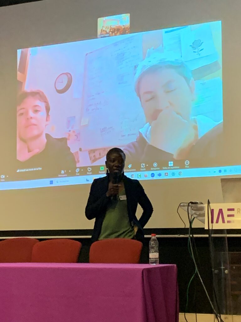 une femme prend la parole, avec un micro, sur l'estrade, avec en son fonds deux femmes en vidéoconférence